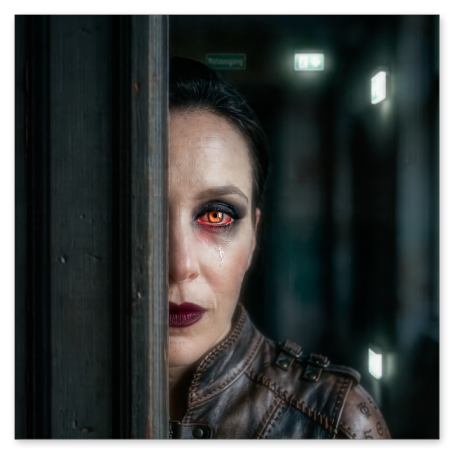 Die linke Gesichtshälfte einer Frau mit einem intensiv rot glühenden Auge und dunklem Make-up ist hinter einem hölzernen Türrahmen sichtbar. Sie trägt eine braune Lederweste mit Schnallen und Tätowierungen auf der Schulter vor einem düsteren Flur.